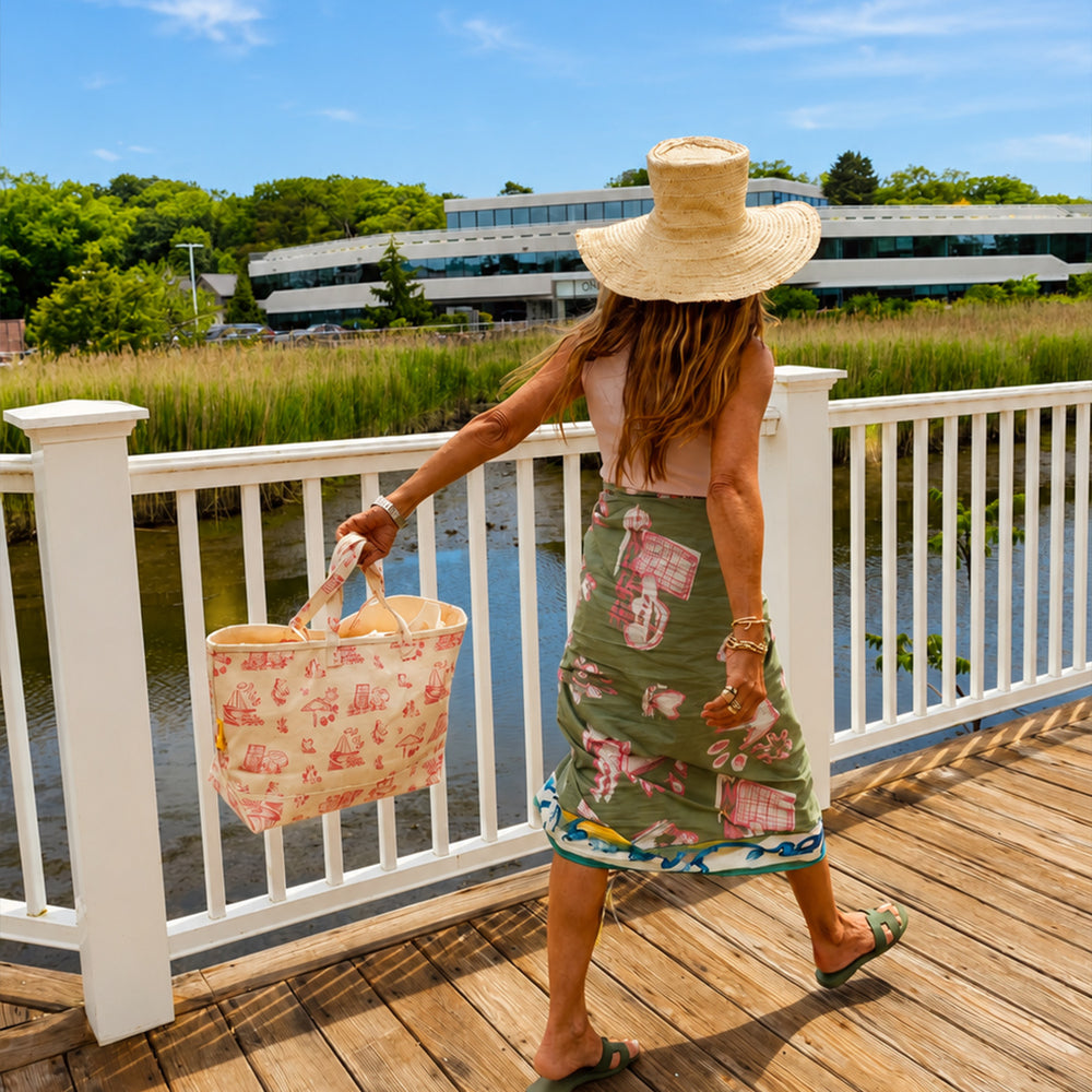 Kerri walking on a sun-lit boardwalk wearing her pink toile sarong wrap skirt, pink KR tank, and wide brim straw hat, carrying a matching pink toile canvas tote bag.