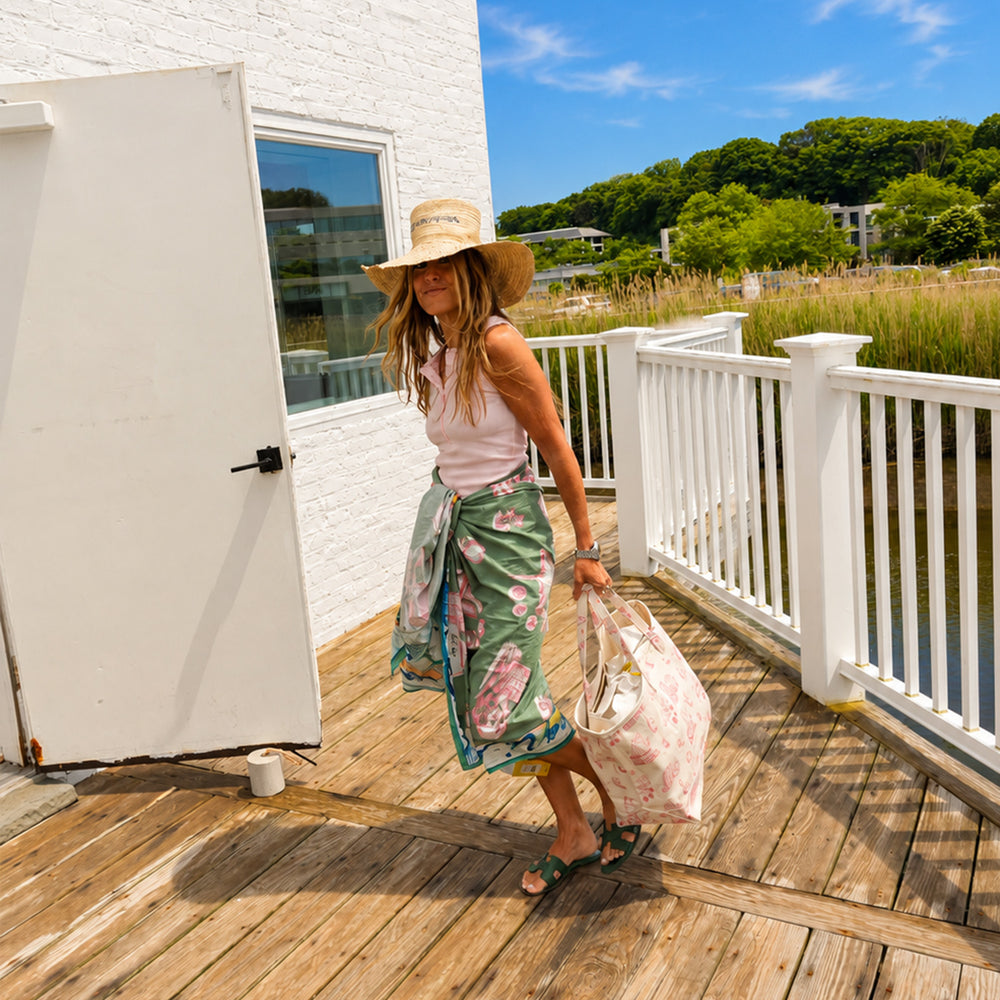 Kerri walking on a sun-lit boardwalk wearing her pink toile sarong wrap skirt, pink KR tank, and wide brim straw hat, carrying a matching pink toile canvas tote bag.