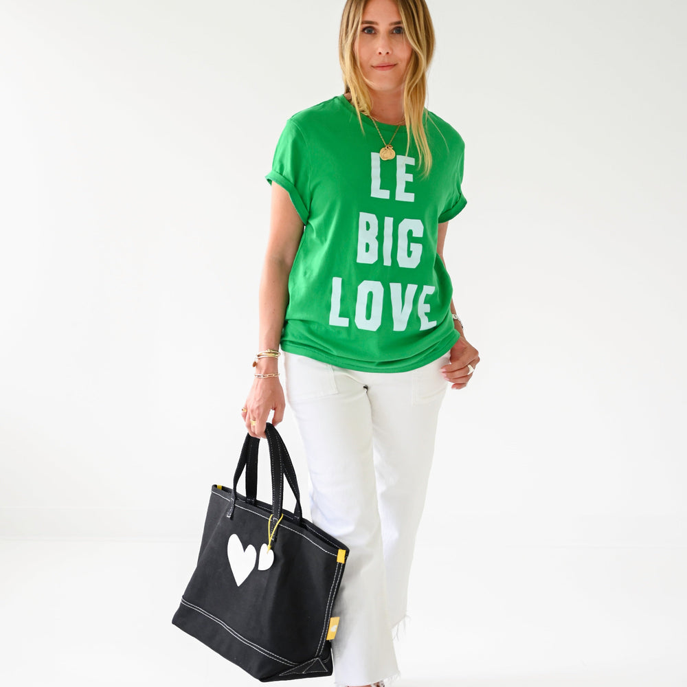 A woman modeling a bright green graphic t-shirt with the words 'BE BIG LOVE' in white, paired with white pants and brown sandals, while holding a black tote bag with a white heart on a plain white studio background.