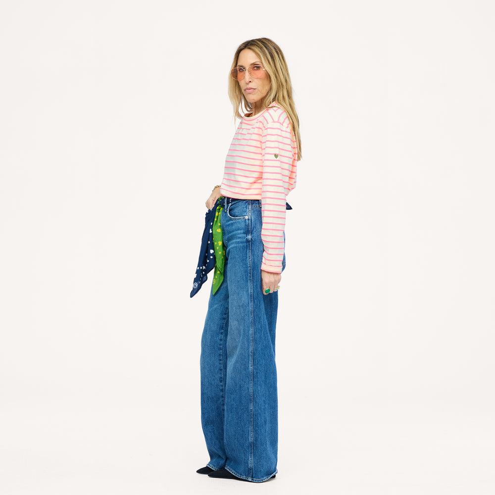 Side view of a woman wearing a Kerri Rosenthal pink and cream striped long sleeve crewneck top with a gold heart detail, styled with wide-leg blue jeans, a green and navy heart print bandana, and pink sunglasses.