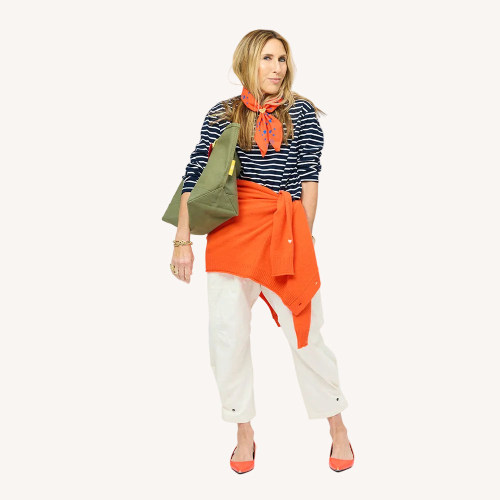 Woman wearing a navy striped top with an orange sweater tied at the waist, an orange bandana, cream pants, orange heels, and an olive bag.