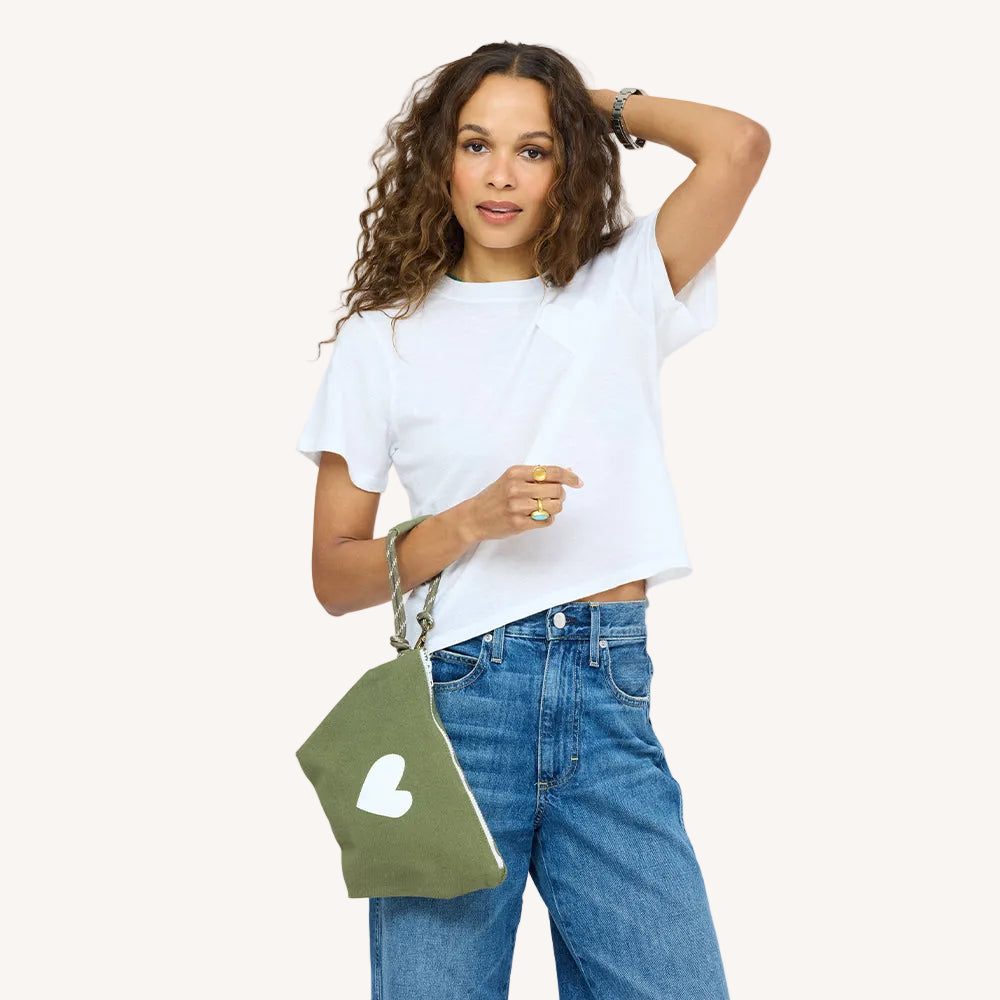 Woman wearing a white t-shirt and jeans, holding an army green heart wristlet pouch.