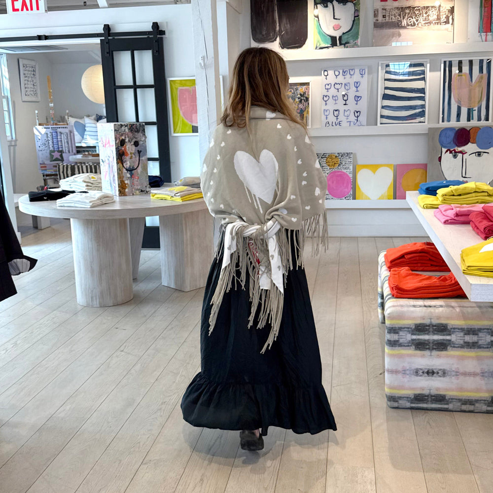Back view of a woman holding up a sage green fringed poncho with a heart pattern and red 'love' stitching, wearing black dream skirt