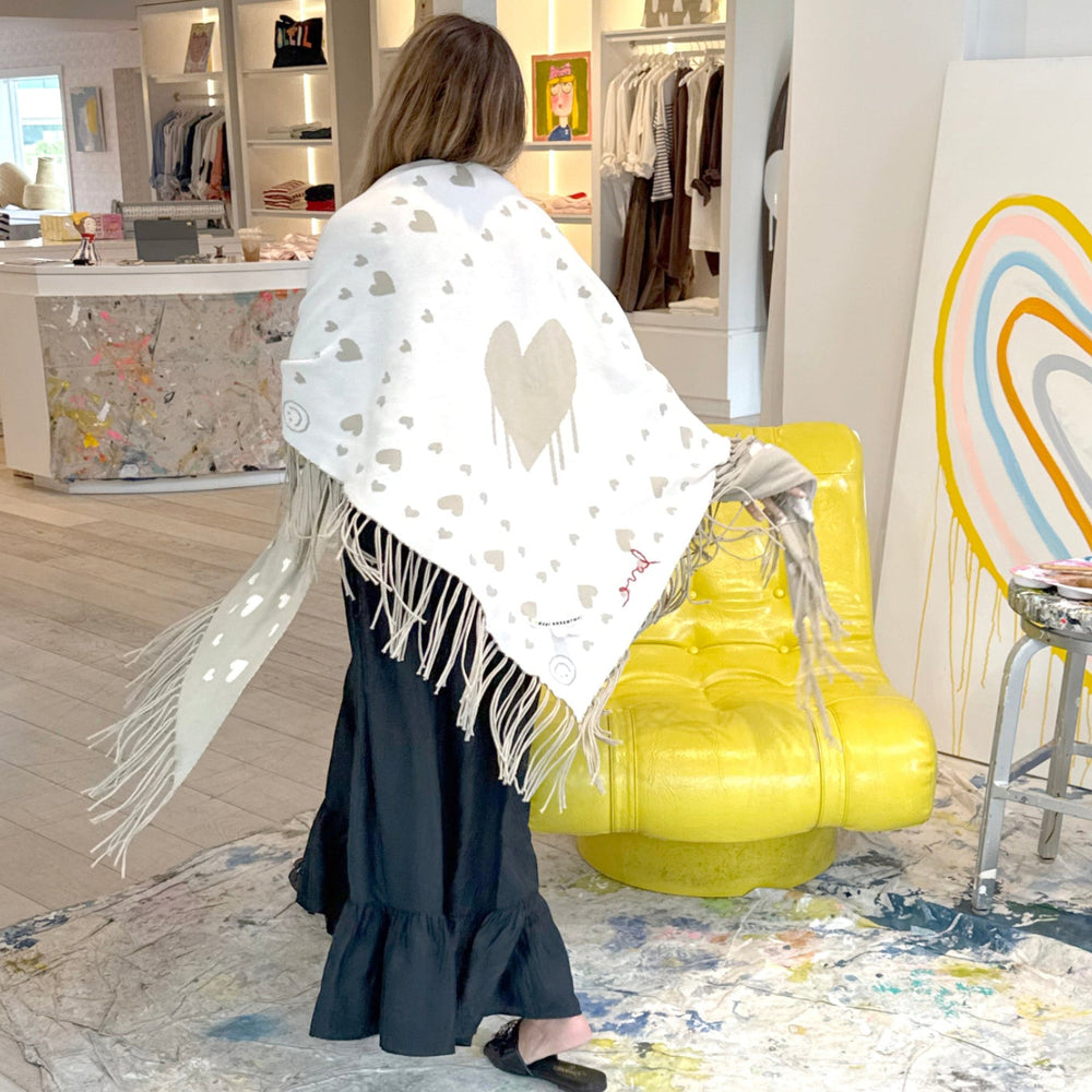 Back view of a woman holding up a cream fringed poncho with a heart pattern and red 'love' stitching, wearing black dream skirt