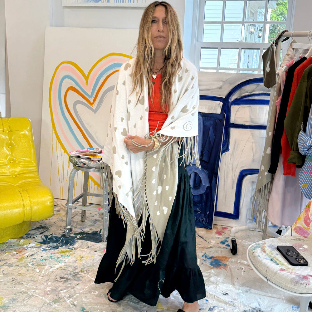 Woman wrapped in a cream fringed poncho with a heart pattern, wearing cream pants, brown boots, and sunglasses.