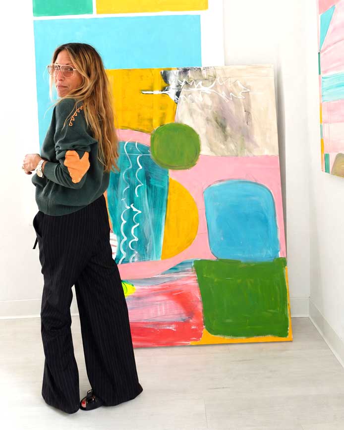 Kerri in a green heart-patch sweater and black pinstripe pants standing next to a colorful abstract painting.
