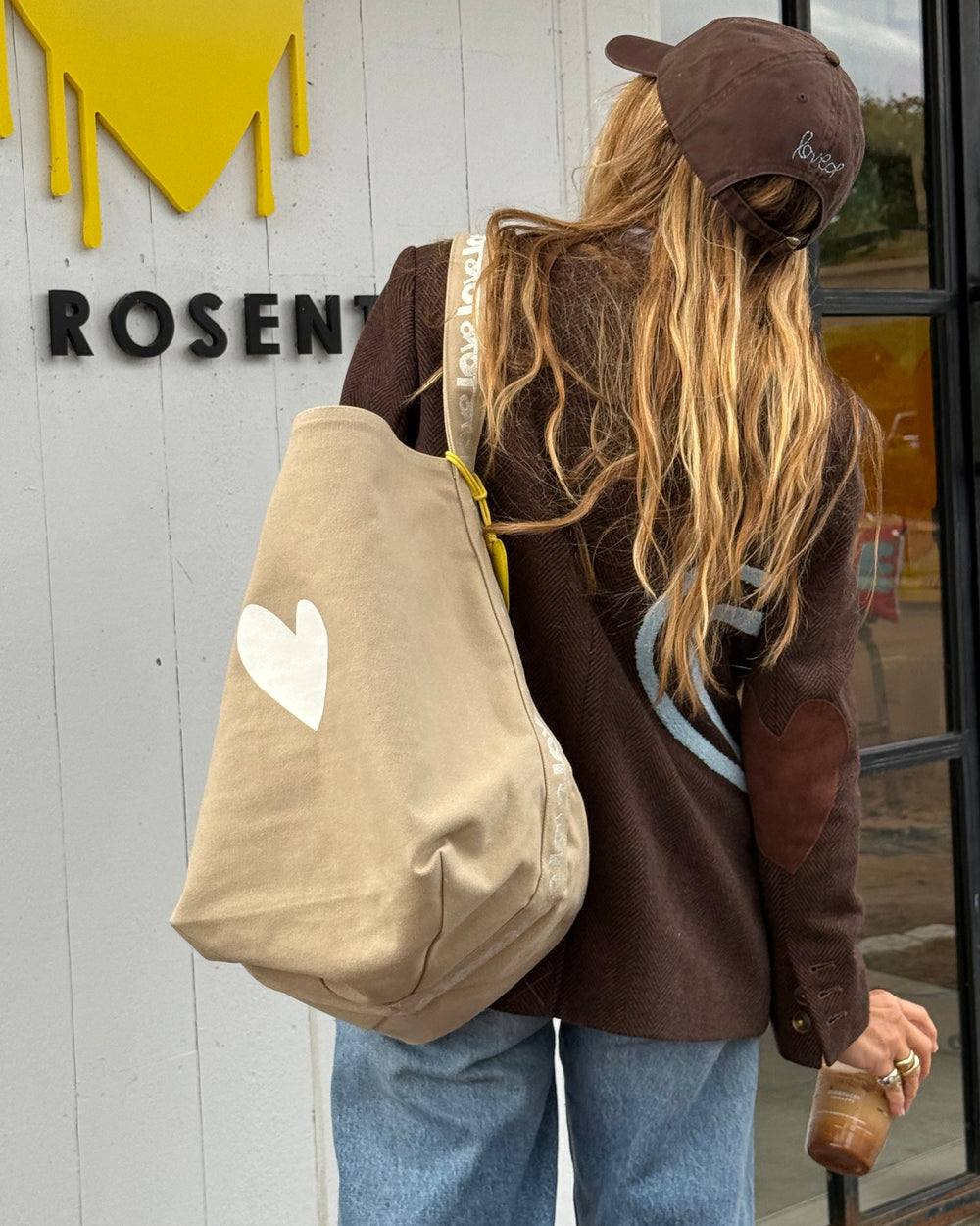 A person from behind wearing a brown cap, brown jacket, and blue jeans, carrying a large beige canvas tote bag with a white heart on it. The background shows a storefront with a white paneled wall, black lettering, and a yellow graphic.