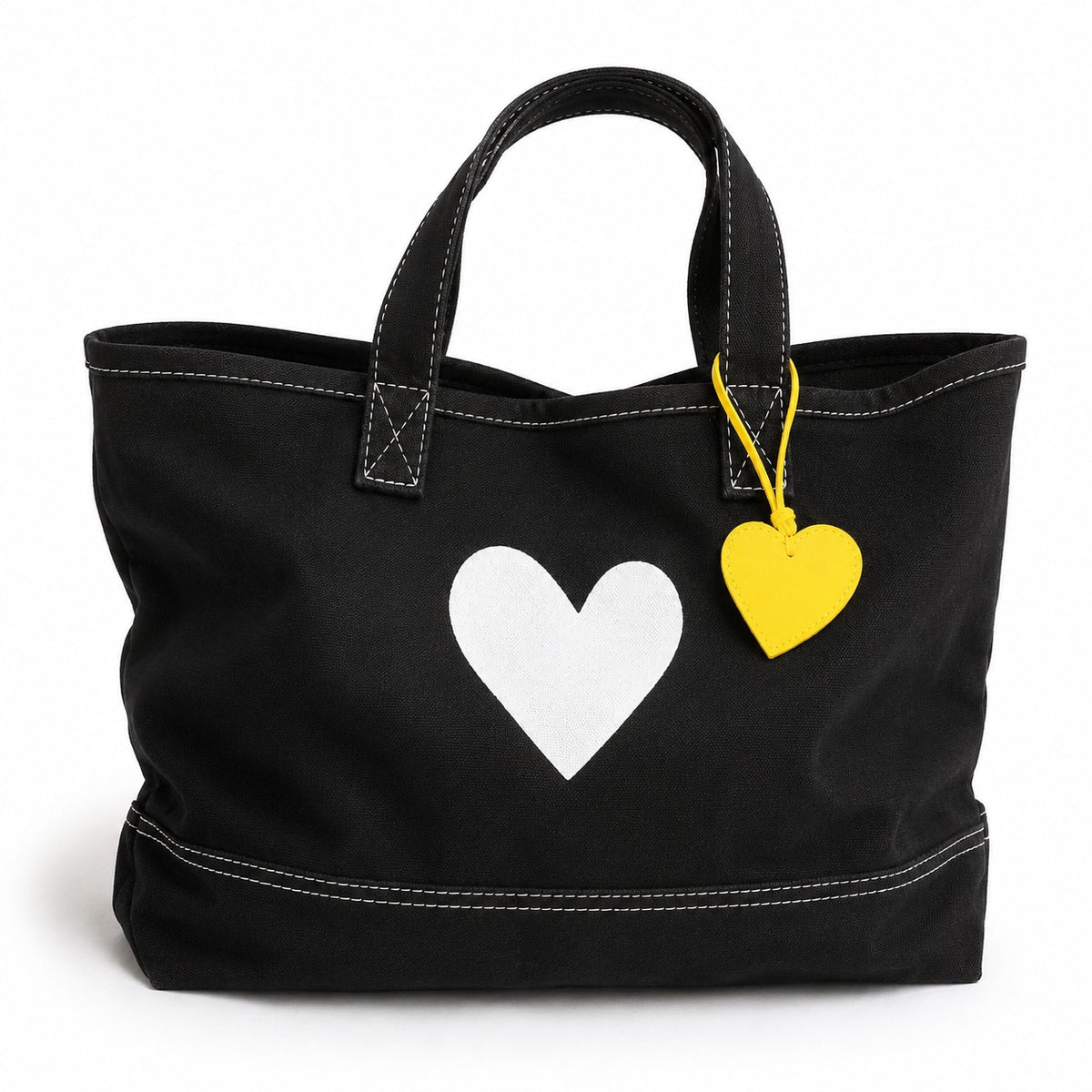 A black canvas tote bag with white contrast stitching, featuring a large white heart on the front and a small yellow heart-shaped tag hanging from a yellow string.