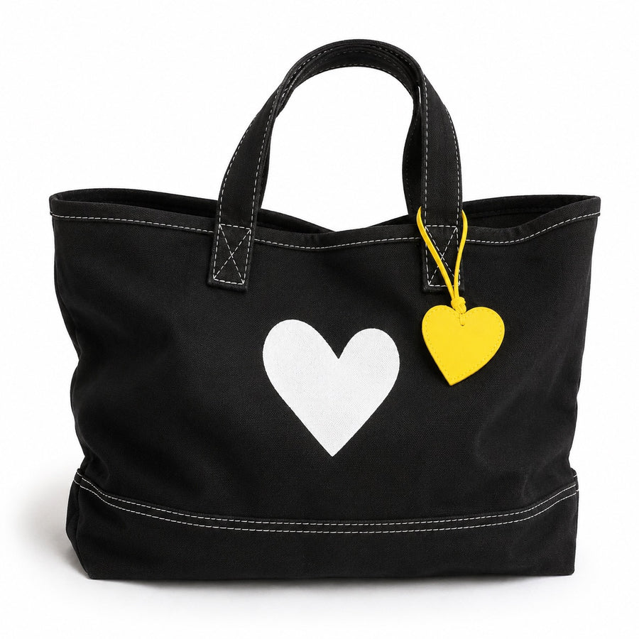 A black canvas tote bag with white contrast stitching, featuring a large white heart on the front and a small yellow heart-shaped tag hanging from a yellow string.