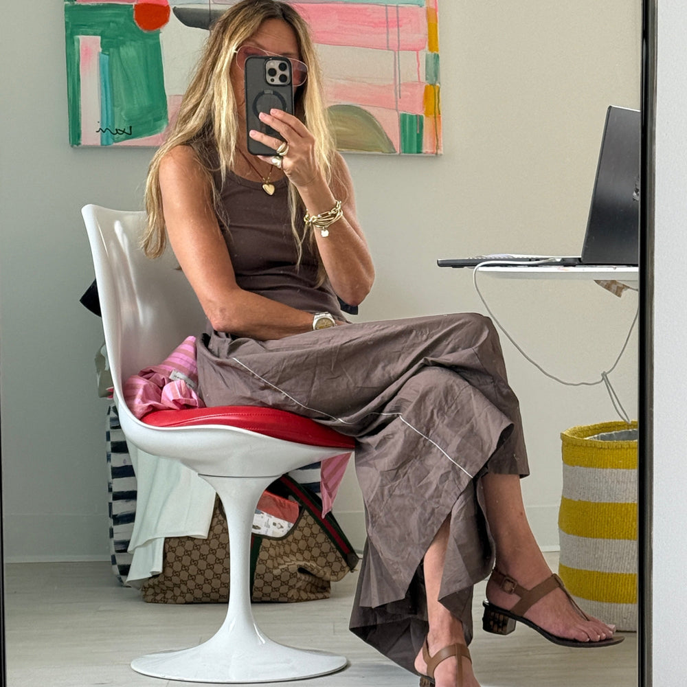 Mirror selfie of a woman seated in a white and red tulip chair, wearing a Kerri Rosenthal brown tank dress with contrast white piping, gold jewelry, and brown thong sandals. Colorful abstract painting hangs on the wall behind her.