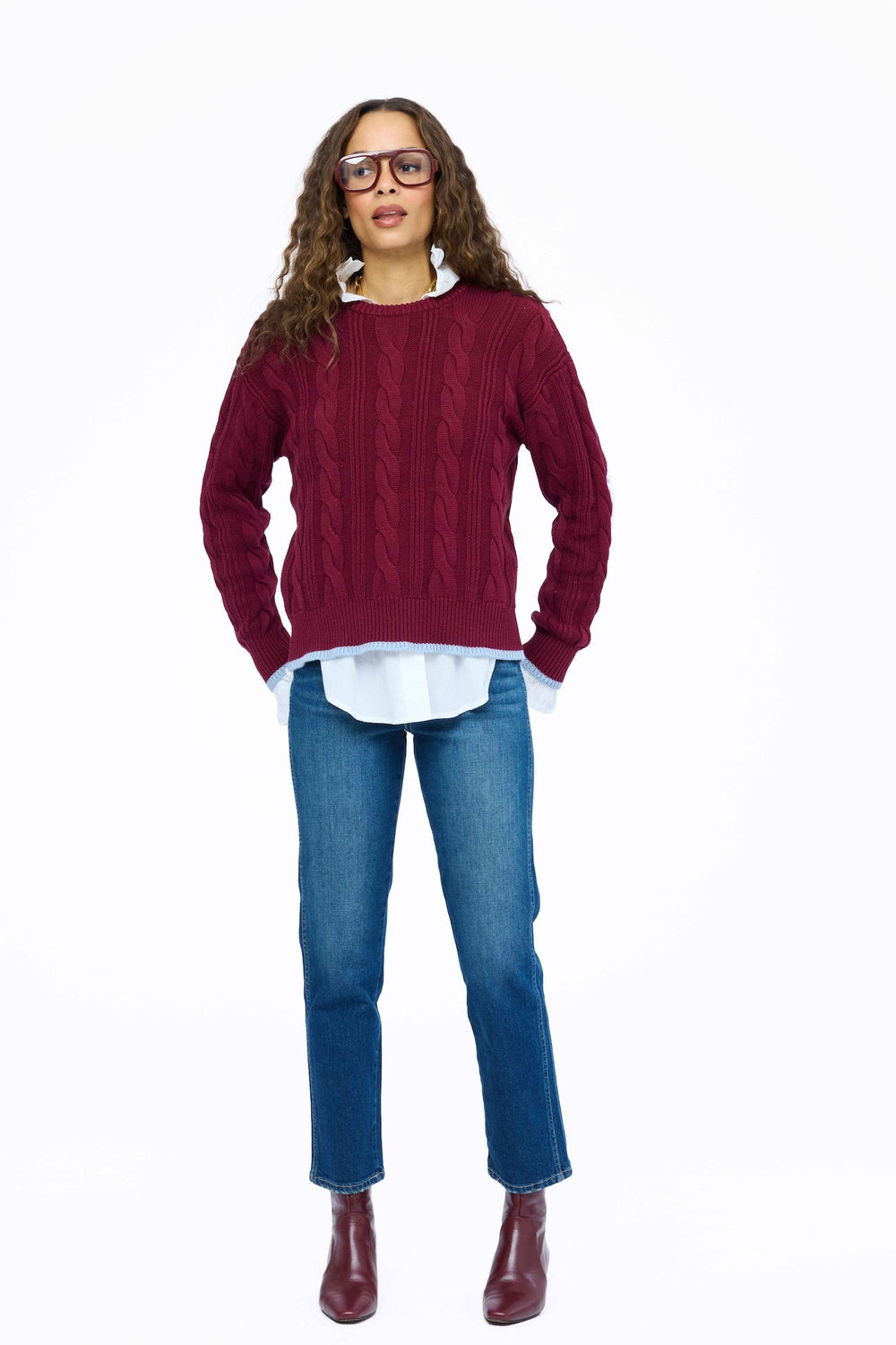 Kerri Rosenthal Maxwork Cable Pullover Sweater in Burgundy, woman standing showing cable knit pattern and contrast patchwork elbows, layered over a white shirt with jeans and burgundy boots.
