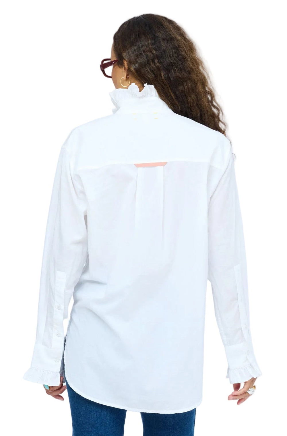 Full-length  back view of model wearing dark red glasses, white ruffle-collar shirt, blue jeans, and burgundy ankle boots, standing with one hand in pocket.