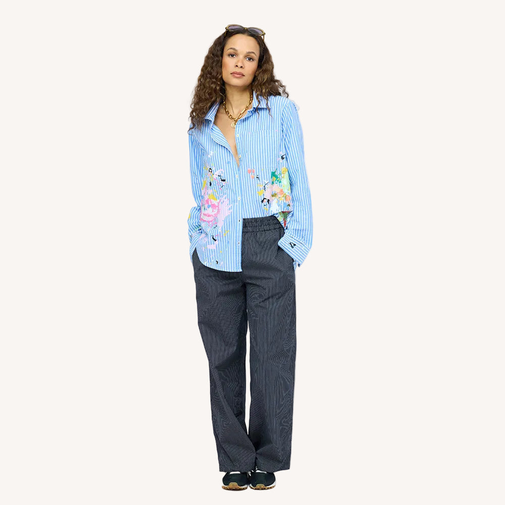 Woman wearing an open blue striped shirt with colorful art print by artist Kerri Rosenthal, navy pinstripe pants, and sneakers.