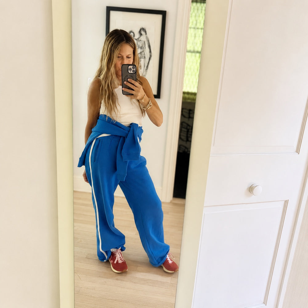 A woman takes a mirror selfie wearing bright blue sweatpants with white side stripes, a white sleeveless top, and red sneakers in a home setting.