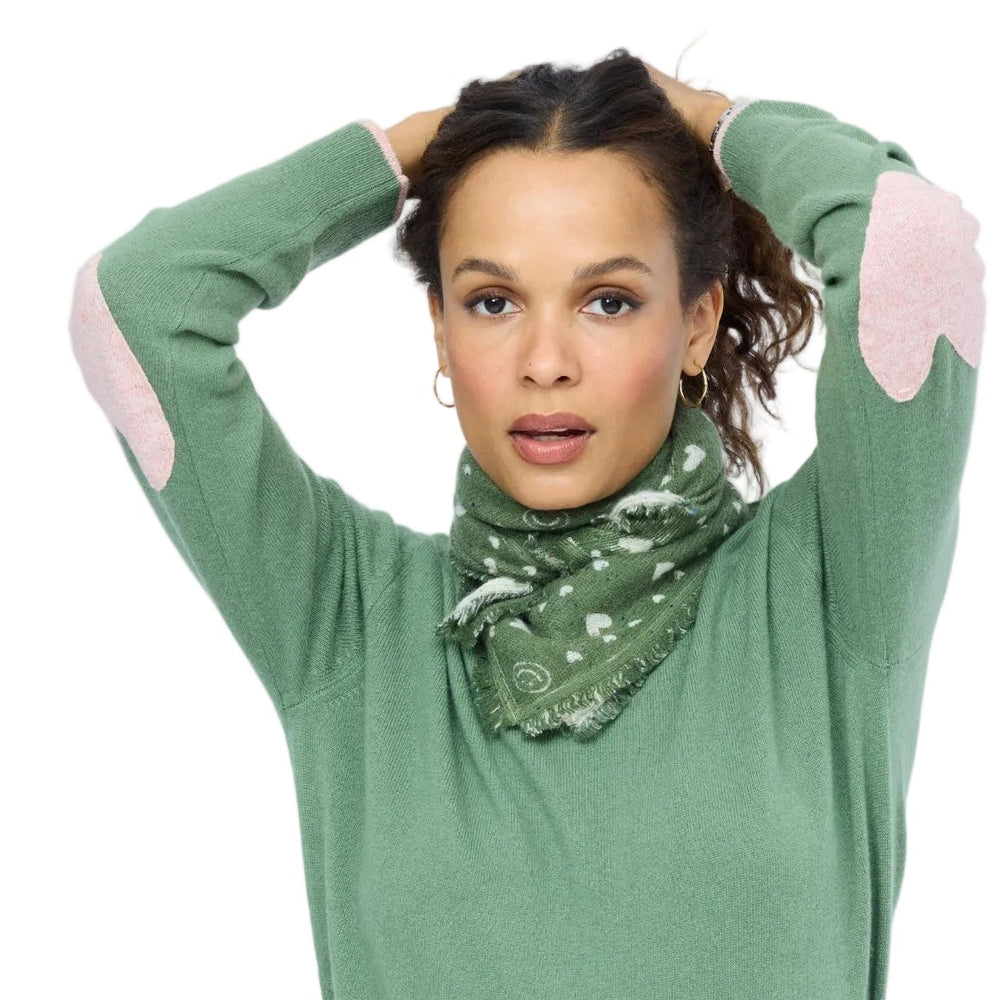 Close-up of model in green patchwork cashmere sweater, arms raised, pink heart patch visible at elbow