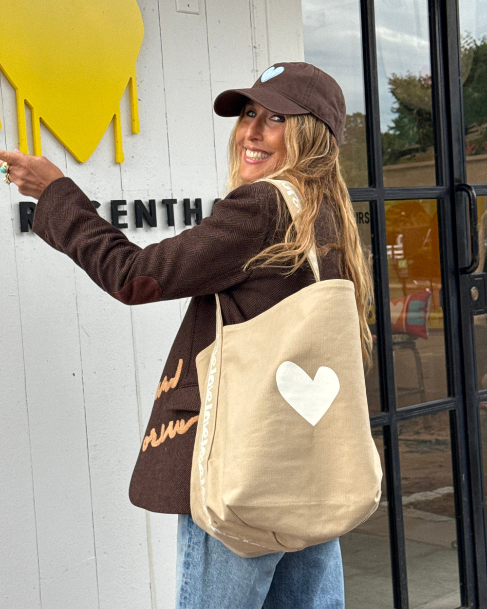 A woman wearing a brown cap and brown jacket stands outside a shop, carrying a beige canvas tote bag with a white heart on the front. She is pointing toward a yellow heart sign on the white wall next to her.