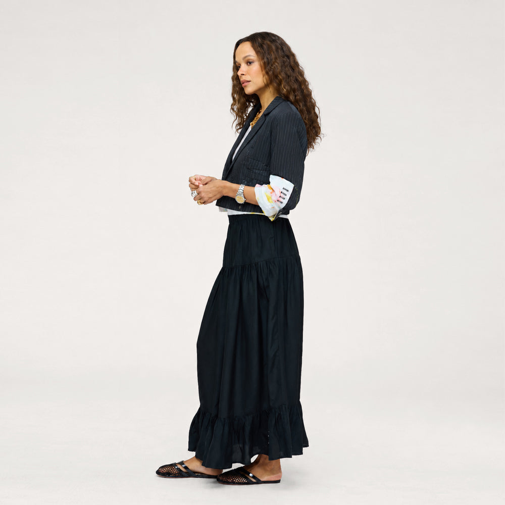 Side view of a woman wearing a Kerri Rosenthal black tiered maxi skirt styled with a black pinstripe blazer, a white art-print shirt, and black woven flat sandals.