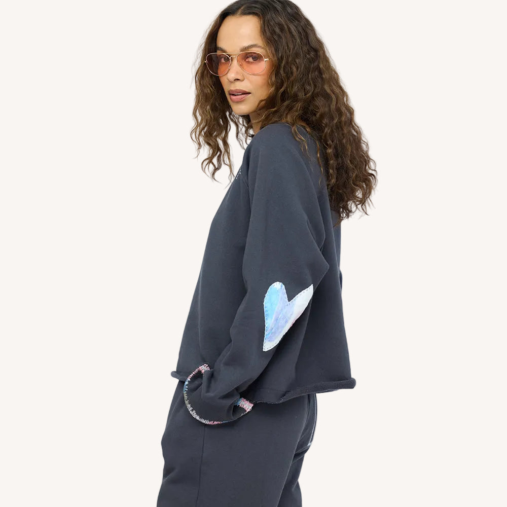 Side view of a woman wearing a charcoal sweatshirt with an paint splat heart elbow patch and sunglasses.