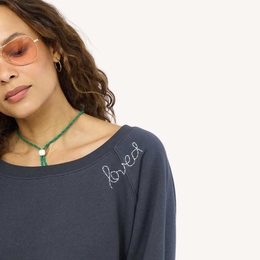 Close-up of a charcoal sweatshirt with 'loved' embroidered on the shoulder, paired with a green cord necklace and sunglasses.