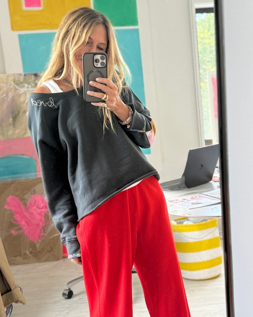 A woman takes a mirror selfie in a room, wearing a dark charcoal off-the-shoulder sweatshirt and bright red wide-leg pants.