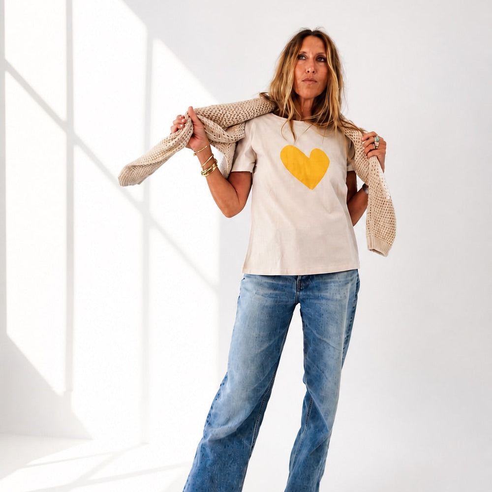 Woman wearing a beige t-shirt with a yellow heart and blue jeans, holding a beige cardigan.