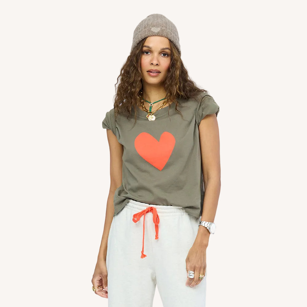 Woman wearing an olive green cap sleeve t-shirt with a large orange heart graphic, paired with a taupe beanie, layered necklaces, and cream sweatpants. Shop Kerri Rosenthal sweats and tees.