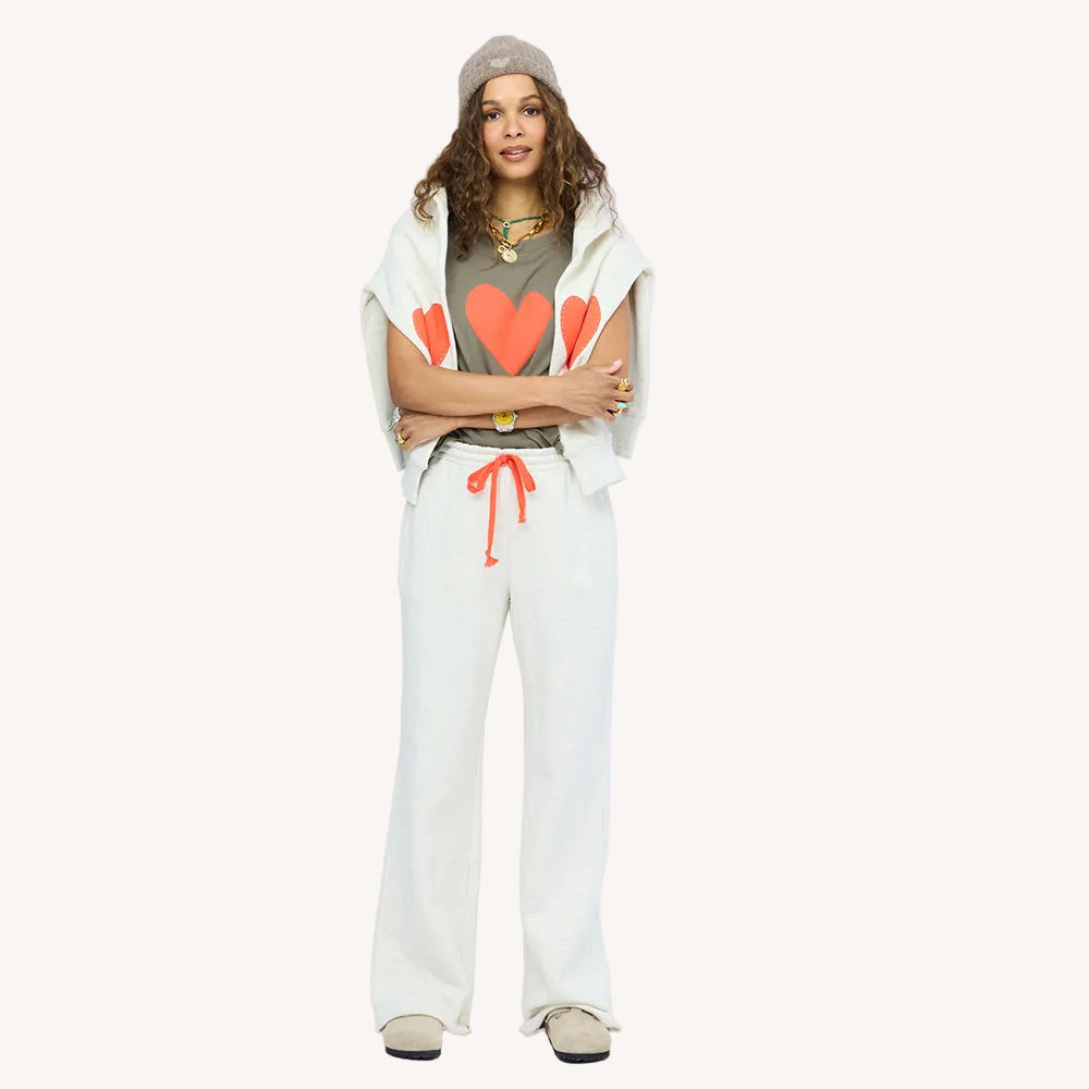 Woman wearing a Kerri Rosenthal olive green heart graphic tee under a cream cropped hooded vest, paired with cream wide leg sweatpants, beanie, and layered necklaces.