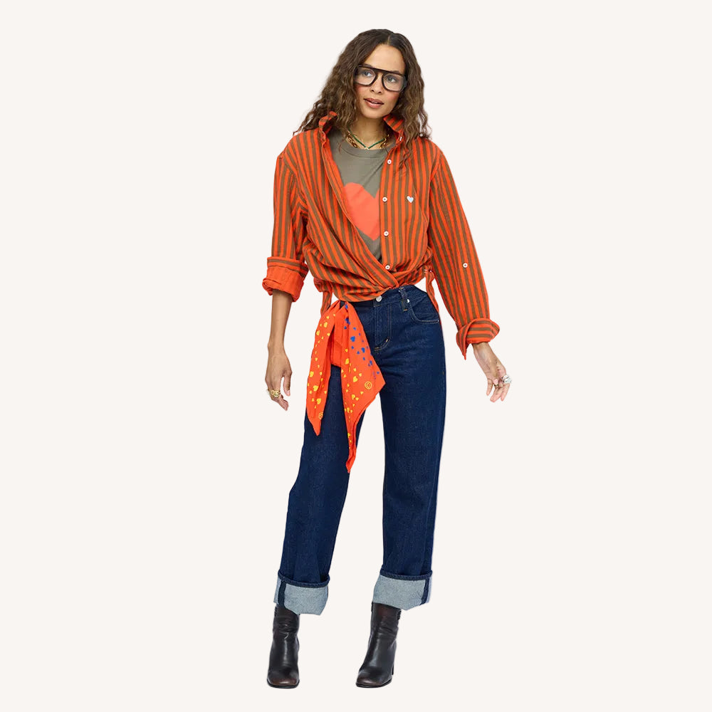Woman wearing an olive green heart graphic t-shirt with an orange printed scarf tied at the waist, paired with dark wash denim jeans and orange and green stripe button down. Shop Kerri Rosenthal