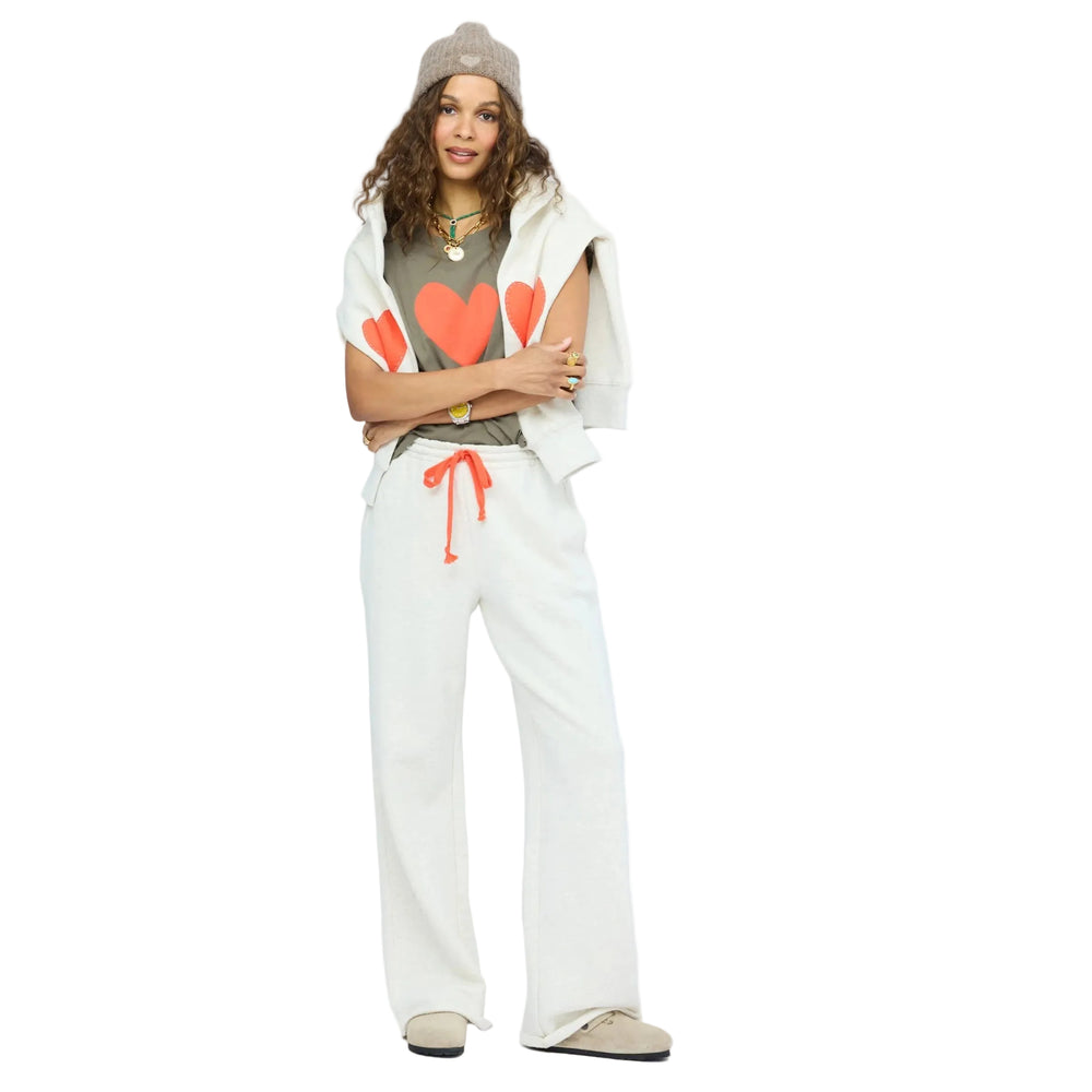 Woman wearing a Kerri Rosenthal olive heart-graphic tank top layered under an oatmeal sweater vest with a coral heart elbow patch, matching oatmeal sweatpants with coral drawstring, a taupe knit beanie, layered gold necklaces, and a wristwatch.
