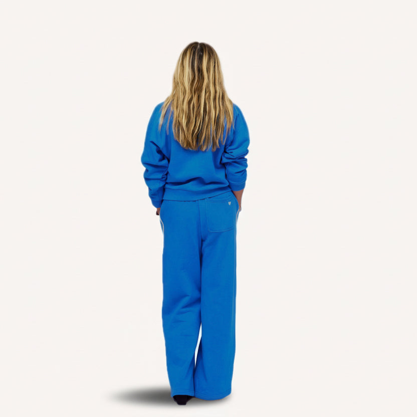 Back view of woman wearing a blue crewneck sweatshirt with and matching sweatpants with a tiny embroidered heart on back.
