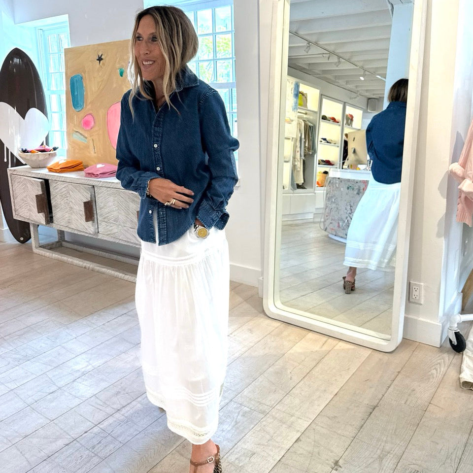 Woman standing in a room with a large mirror, wearing a denim jacket and white pants.