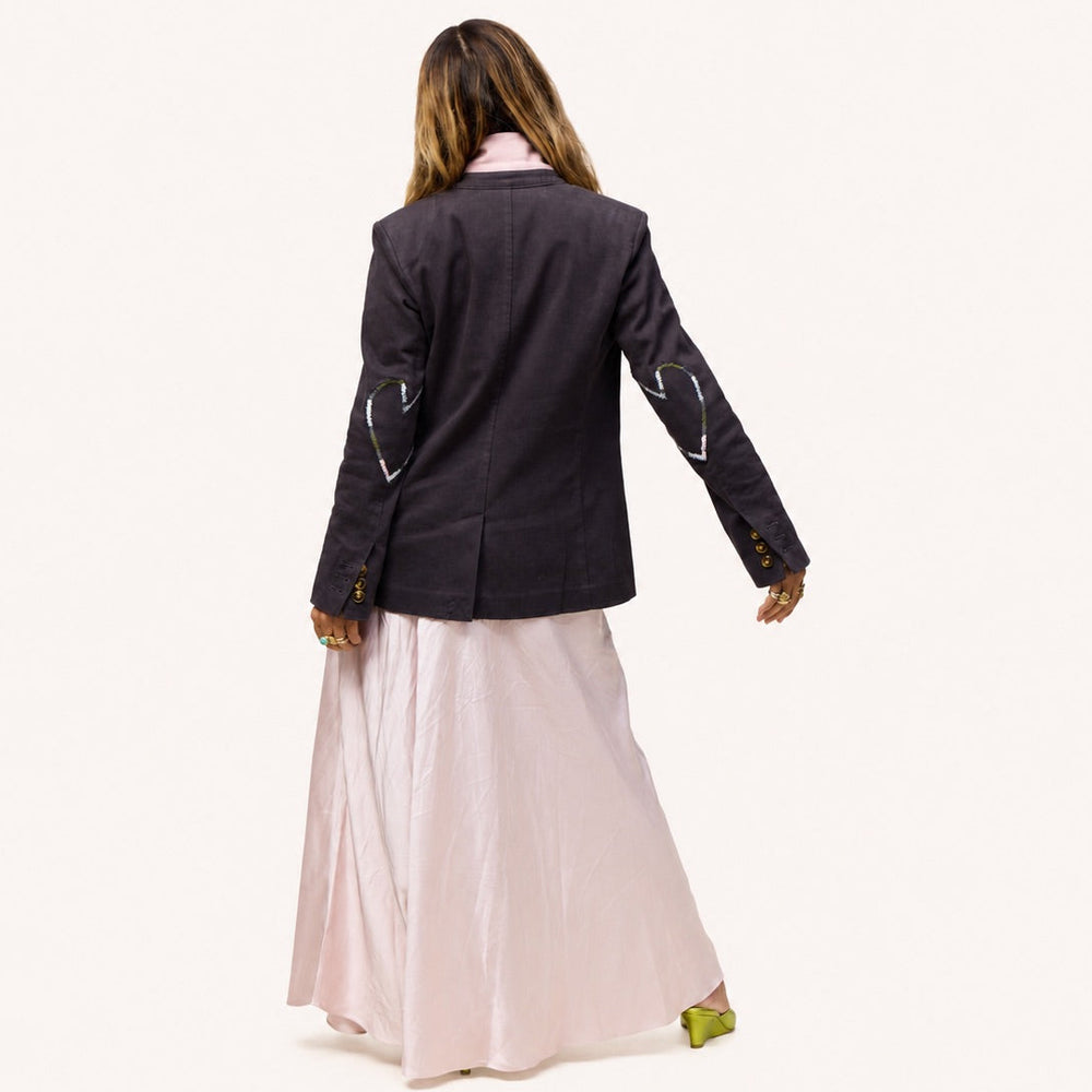 Back view of the Workday Blazer in Anthracite, a structured brown-grey blazer with heart-shaped stitched patches on both sleeves, worn by a woman in a pink maxi skirt. Kerri Rosenthal women's blazers