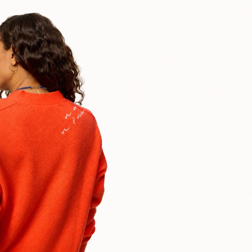 Back view of an orange knit sweater with embroidered cursive script detail on the upper shoulder.