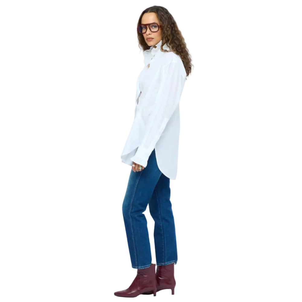 Side view of model with curly brown hair  wearing dark red rectangular glasses, layered gold necklaces, a Kerri Rosenthal white ruffle-collar button-down shirt, and blue jeans, hand tucked in pocket.