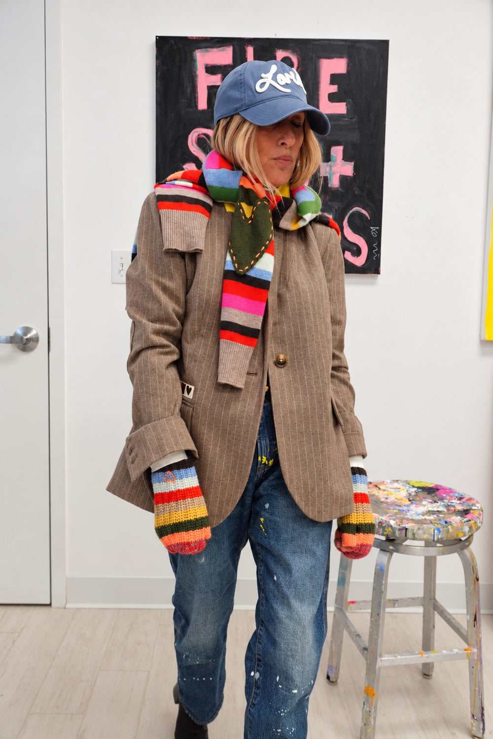 Person wearing the Kisses Mittens in multicolor stripe with the Fireside Scarf, the Loved Baseball Hat, and the Workday Love Brigade Blazer in taupe, standing by a stool