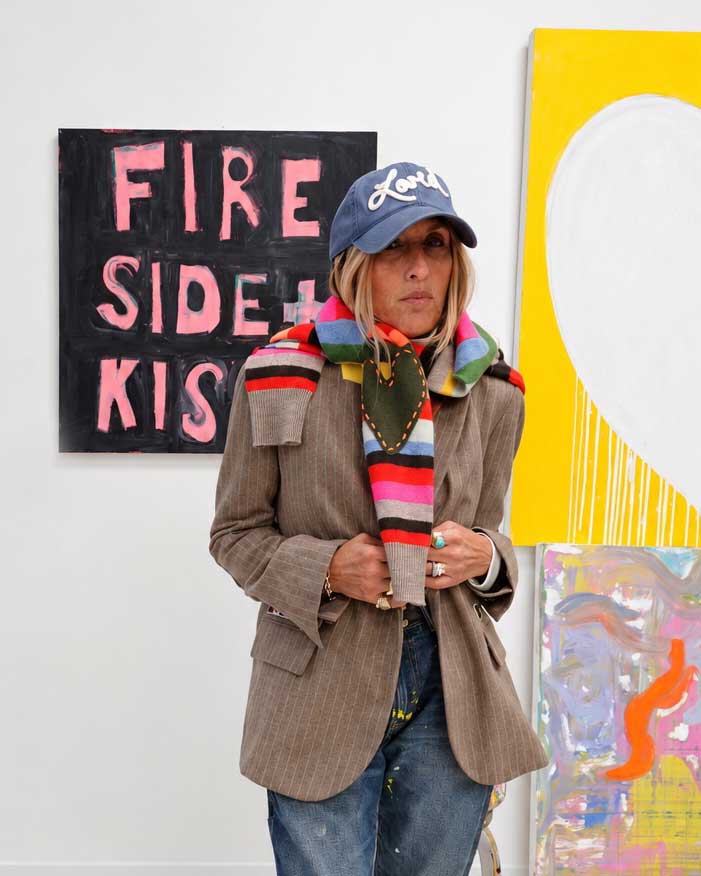 Person wearing the Workday Love Brigade Blazer in taupe with the Loved Baseball Hat, the Fireside Scarf, and Kisses Mittens in multicolor stripe, standing in front of artwork