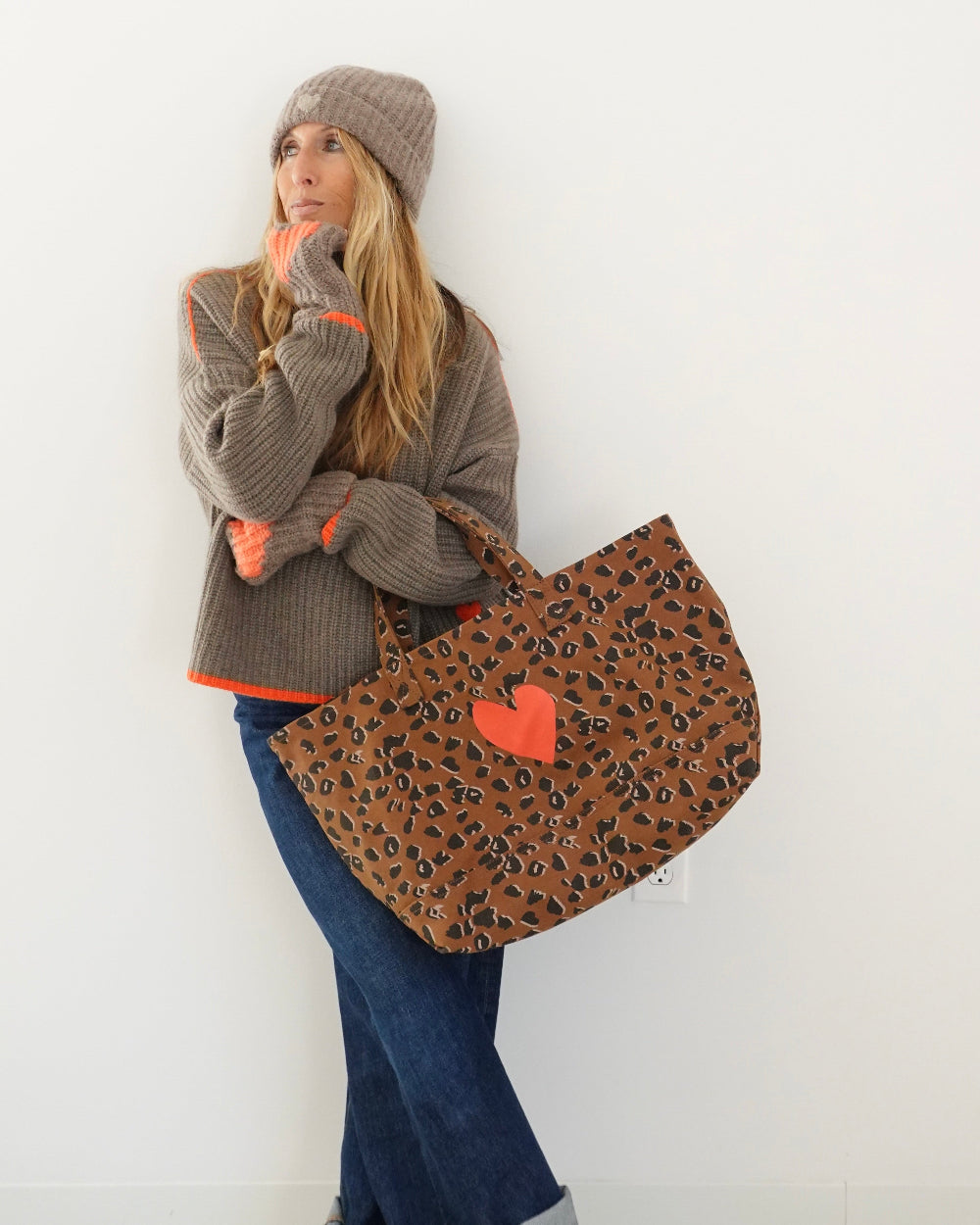 A woman wearing a brown knit beanie, gray knit sweater, and blue jeans stands against a white wall holding a large leopard-print tote bag with a bright orange heart on the front.