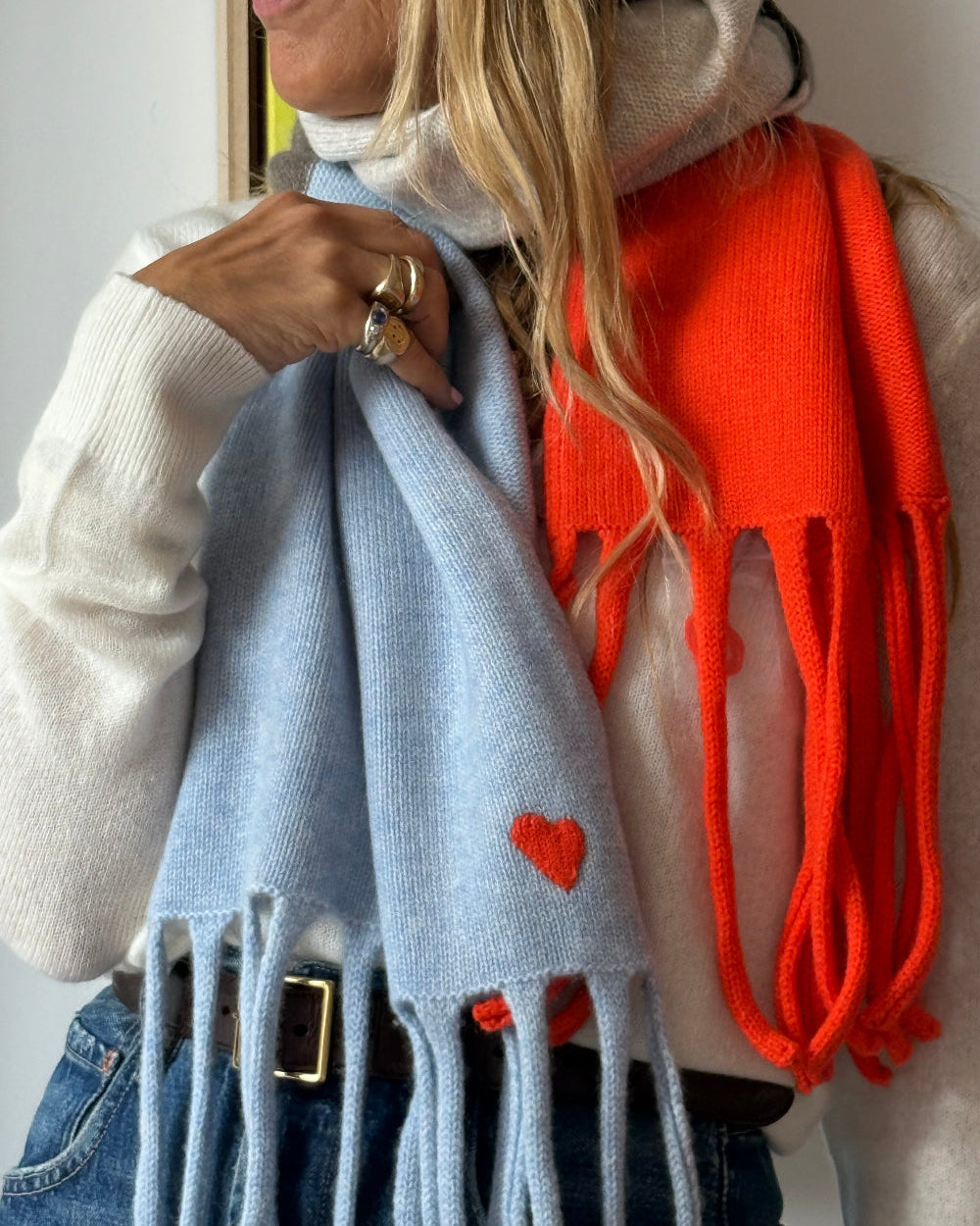 A woman wearing a light blue knit scarf with a small red heart and fringe, layered over a white turtleneck sweater and blue jeans, with a bright orange knit scarf draped over her shoulder.