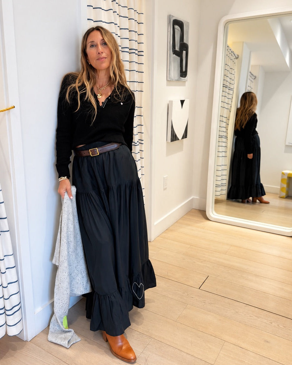 A woman stands in a boutique wearing a black long-sleeve top and a long black skirt with a brown belt, holding a light gray cardigan. The boutique has white walls, a large mirror, patterned curtains, and light wood floors.