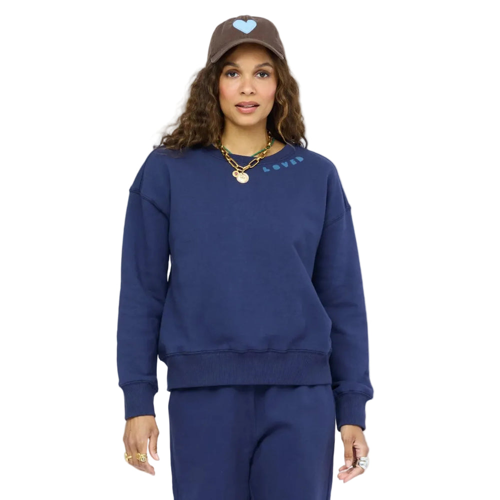 Woman facing forward wearing a navy blue Kerri Rosenthal sweatshirt with 'LOVED' text and a light blue heart graphic, matching navy wide-leg sweatpants, taupe slip-on shoes, a brown baseball cap with a heart design, and layered gold jewelry.