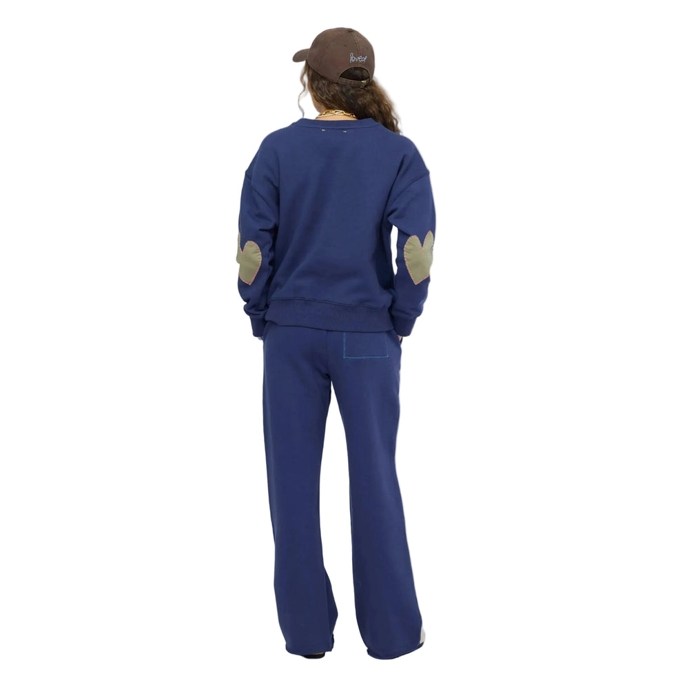 Back view of woman wearing a navy blue sweatshirt with contrast elbow patches, matching navy sweatpants, taupe slip-on shoes, and a brown baseball cap with a light blue heart, gold layered necklaces. Shop Kerri Rosenthal sweats.