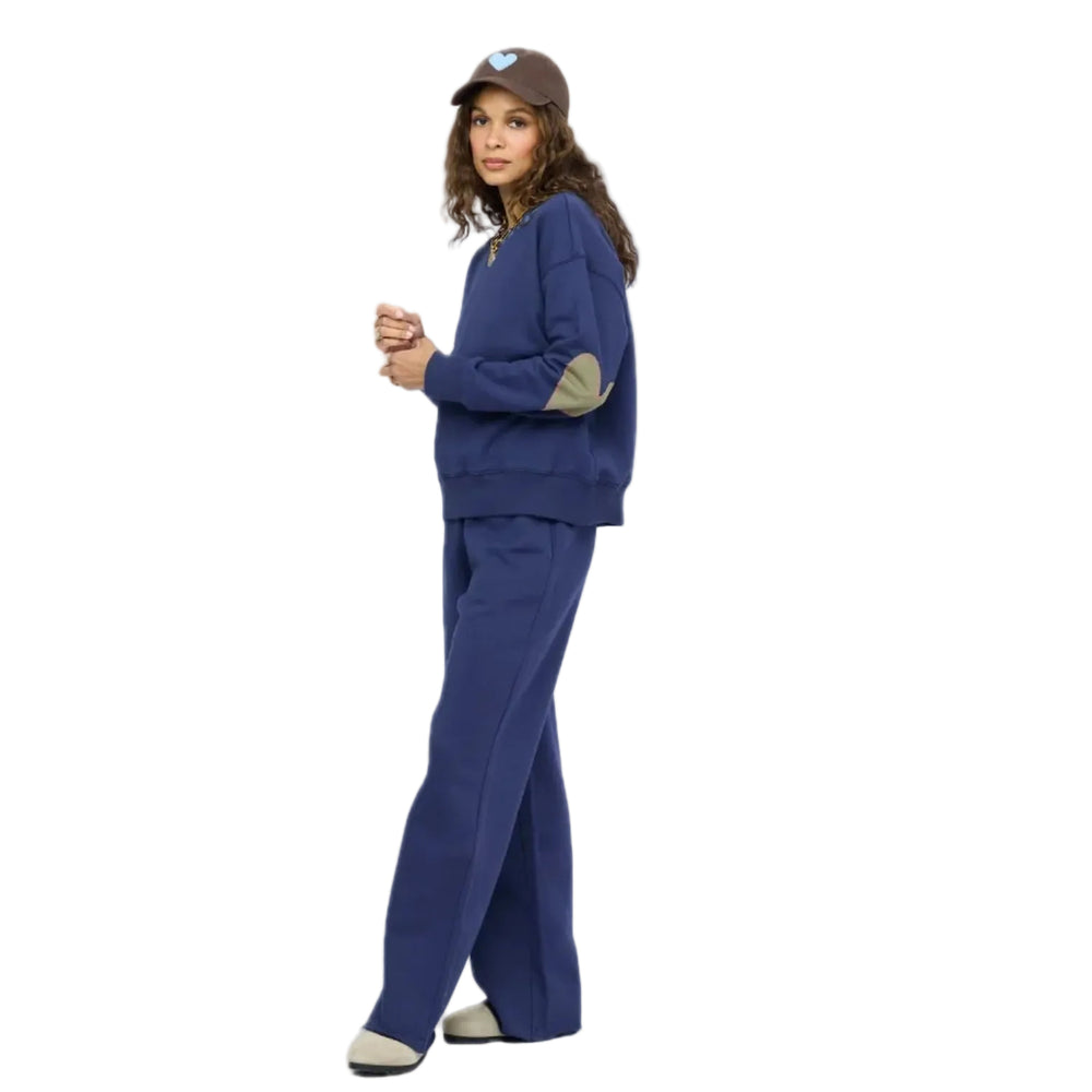 Woman in profile wearing a navy blue sweatshirt with contrast elbow patches, matching navy sweatpants, taupe slip-on shoes, and a brown baseball cap with a light blue heart, gold layered necklaces. Shop Kerri Rosenthal sweats.