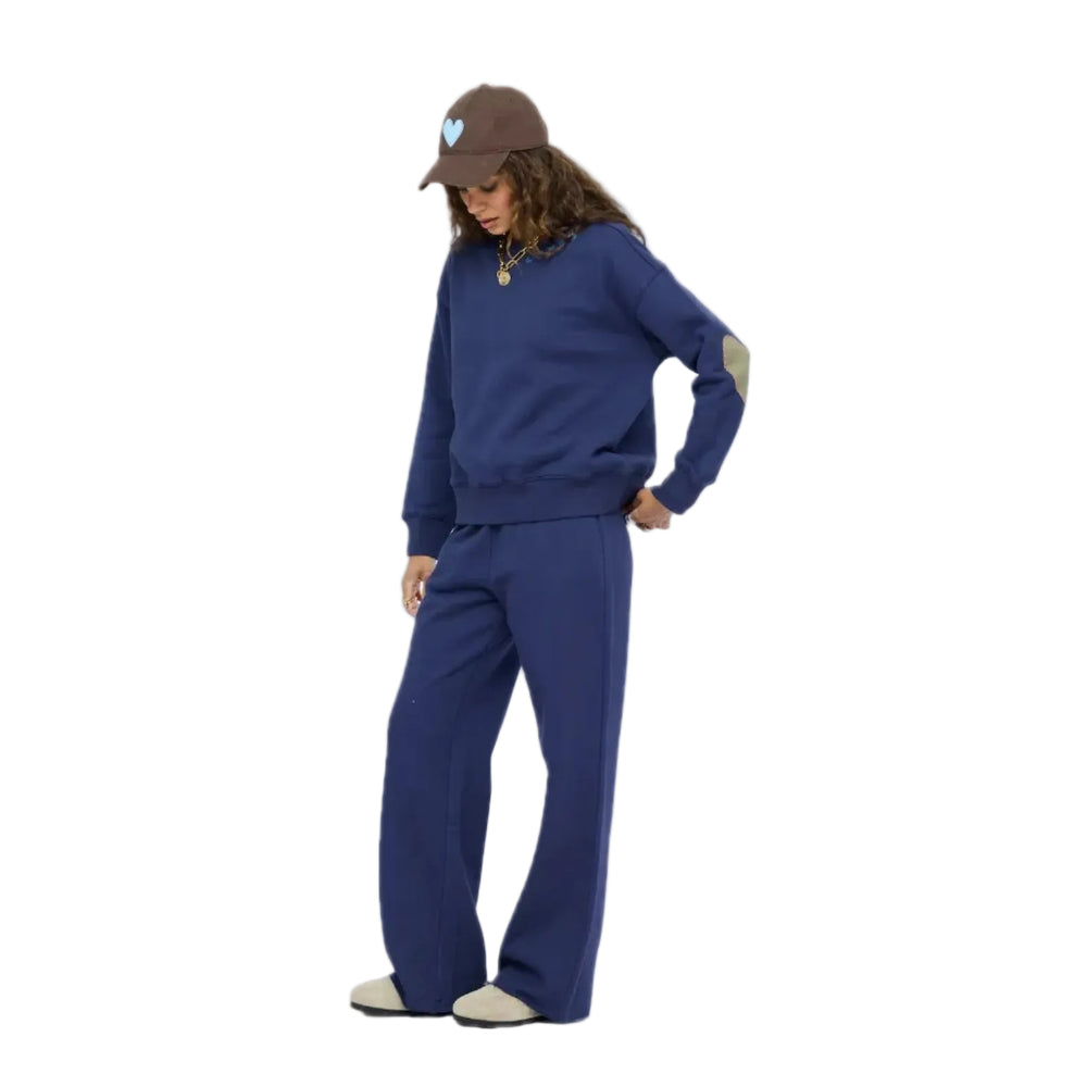 Woman in profile wearing a navy blue sweatshirt with contrast elbow patches, matching navy sweatpants, taupe slip-on shoes, and a brown baseball cap with a light blue heart, gold layered necklaces. Shop Kerri Rosenthal sweats.