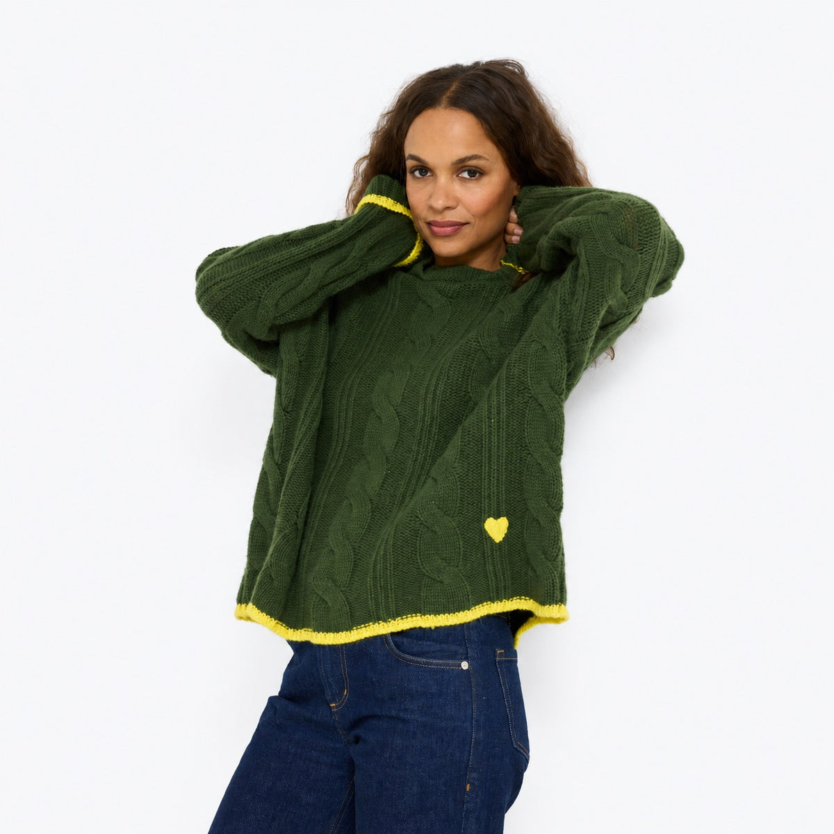 Model wearing the Hearth Sweater in spruce with yellow tipped cuffs and hem and a small yellow heart, hands behind her head, front view
