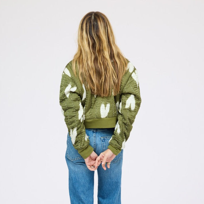 Close-up of woman wearing an olive green quilted bomber jacket with heart print, jeans, and black heels. Shop jackets and blazers at Kerri Rosenthal.