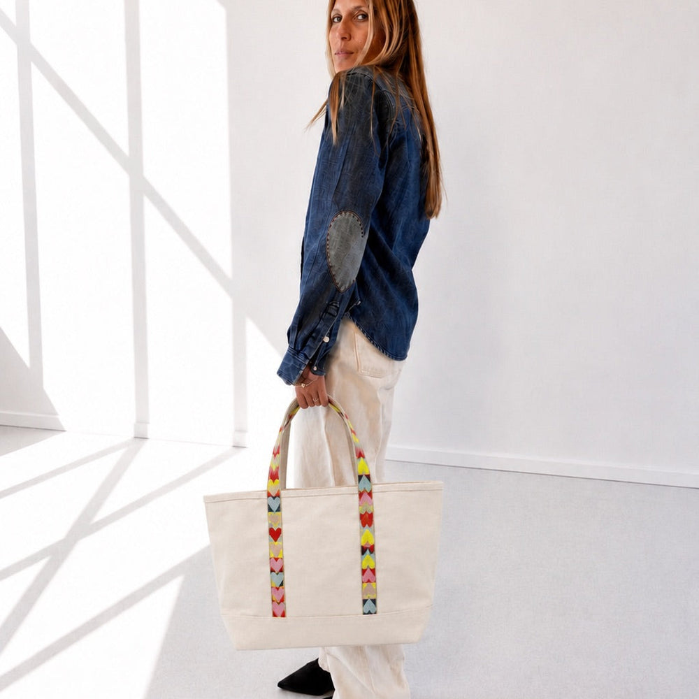 A woman stands in a bright white room holding a cream canvas tote bag with colorful striped handles. She is wearing a blue denim jacket, white pants, and black shoes.