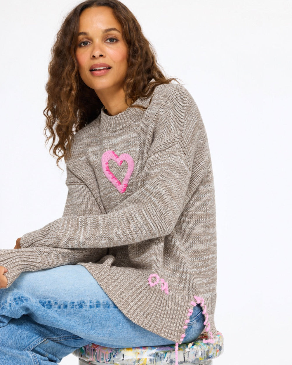 Model seated on a stool wearing the Lovey Heart Pullover in taupe with jeans
