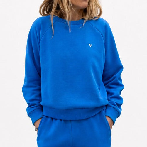 Woman wearing a blue crewneck sweatshirt with a small heart logo and matching sweatpants.
