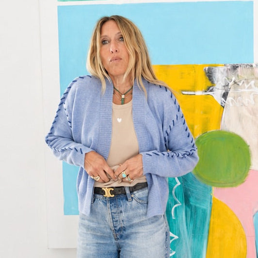 Model wearing the Kerri Rosenthal blue cardigan with whipstitch trim, open over a white top, hands in dark denim jean pockets, layered necklaces.
