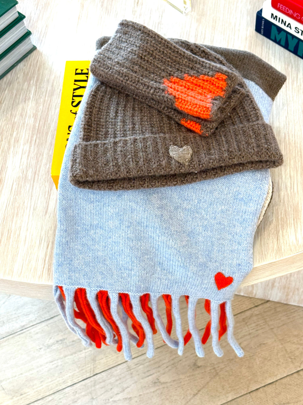 A light blue knit scarf with red and white fringe, folded with a brown knit beanie that has an orange heart and a small silver heart charm, displayed on a light wood surface.